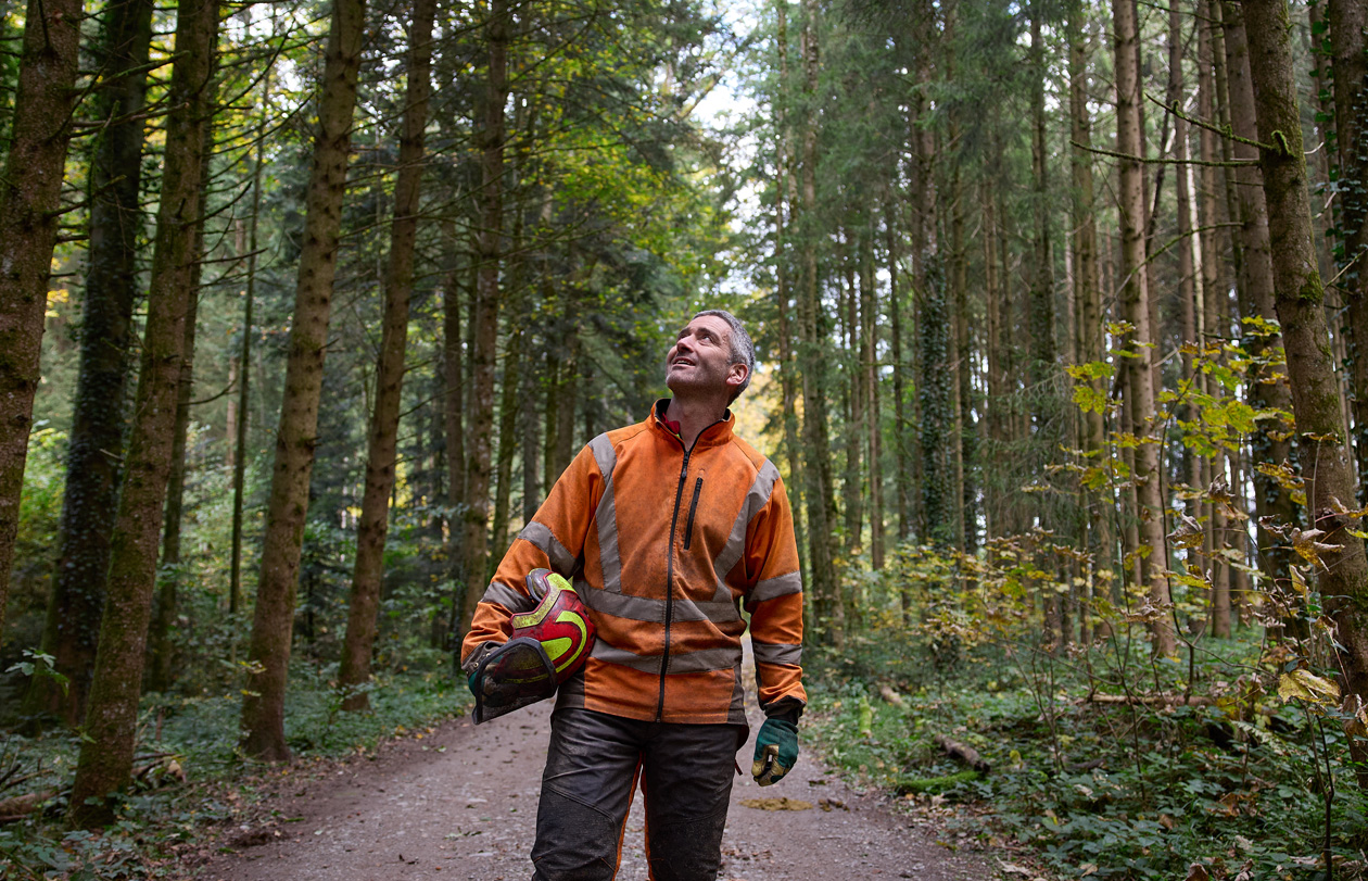 Man wearing an orange high-visibility jacket holding a hard hat while standing on a forest path in a sustainably managed forest in Switzerland