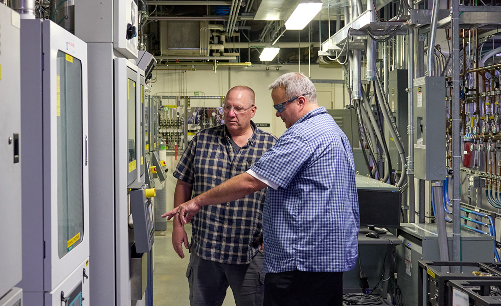 Two people wearing safety glasses talk near facility equipment