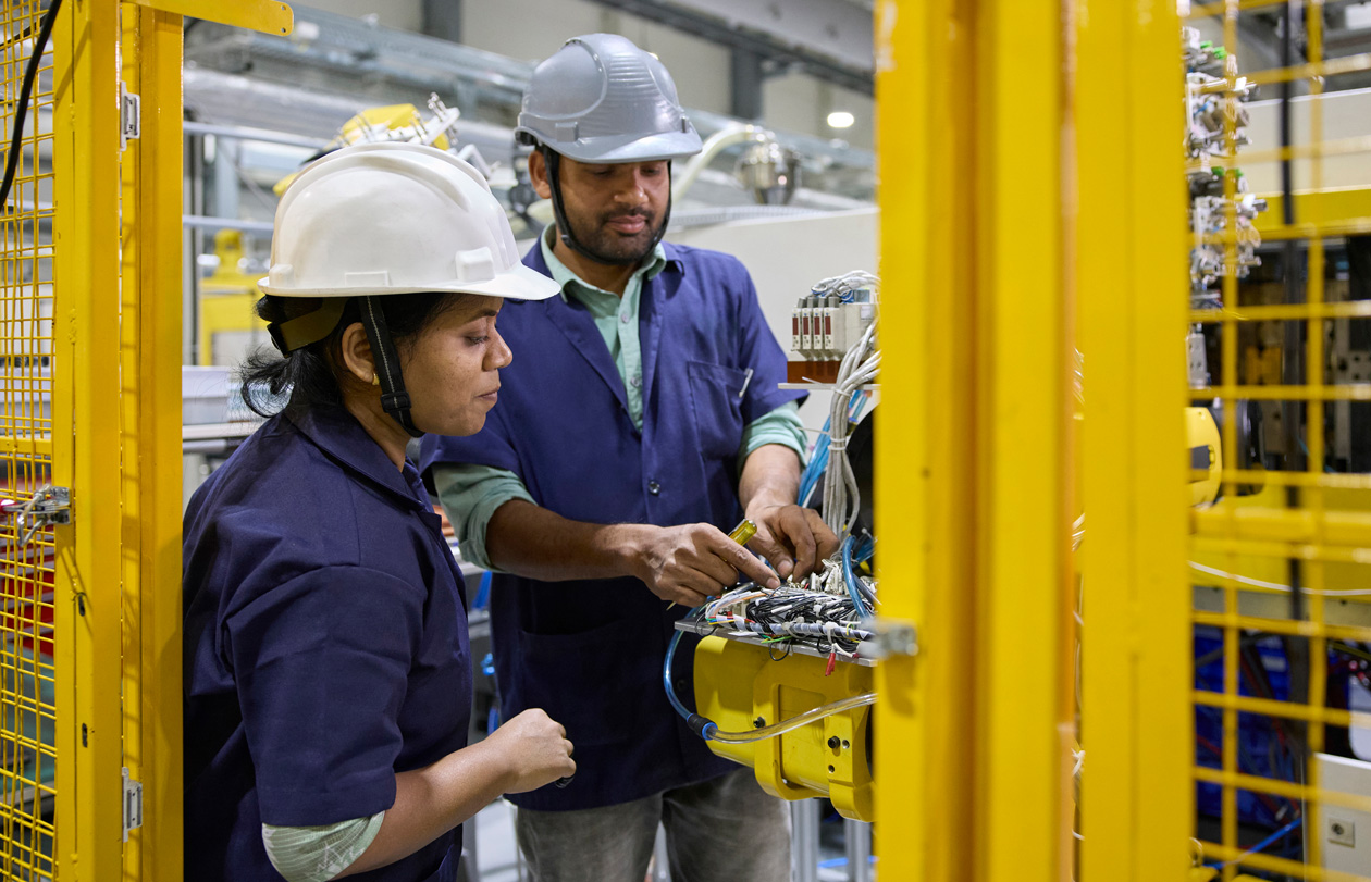 Two employees in work smocks and hats stand at their work stations operating assembly machines